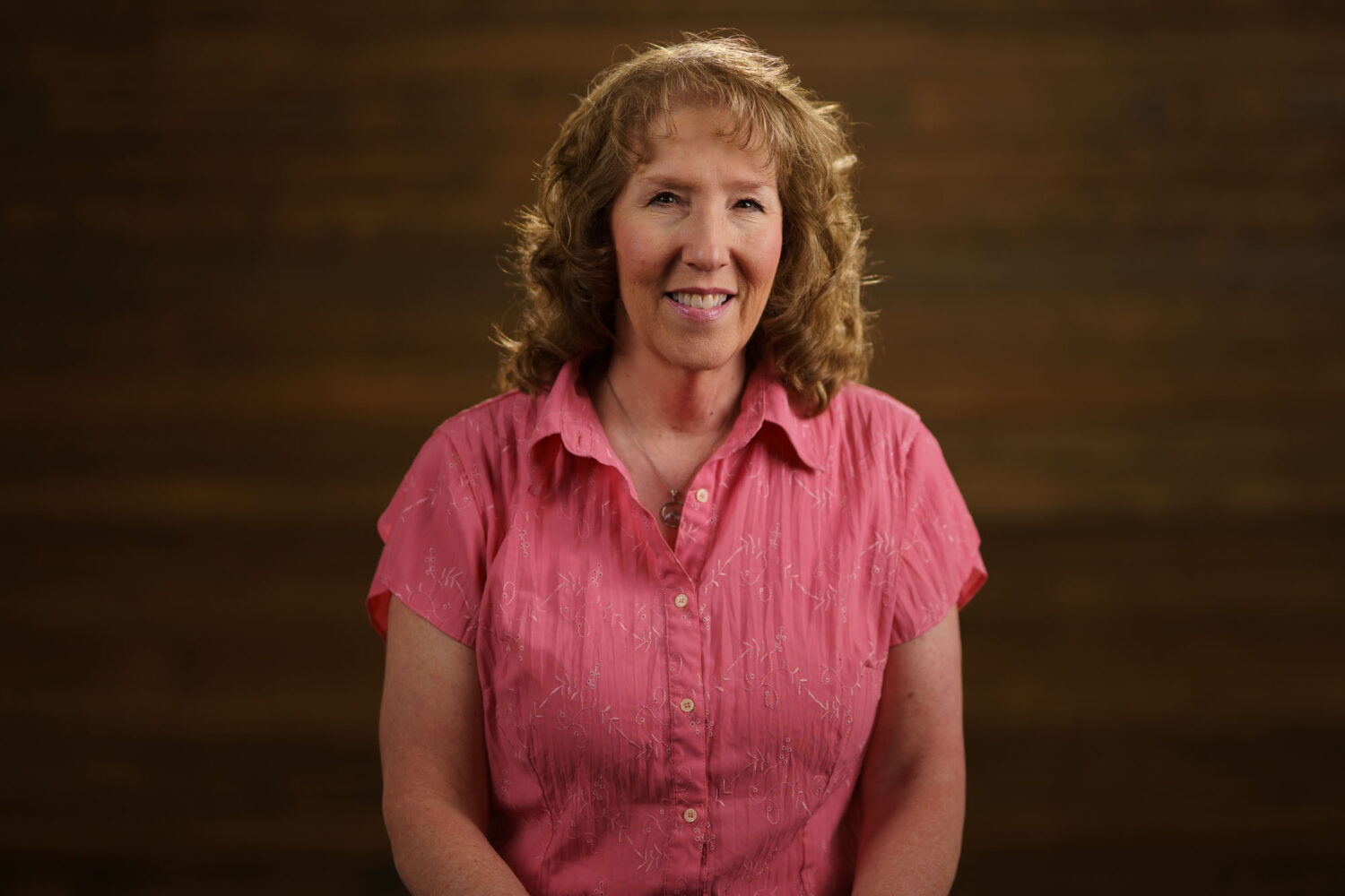 A woman in a pink shirt smiling in front of a wooden background. profile picture