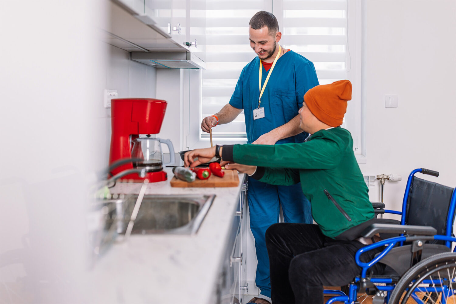 A man in a wheelchair cuts vegetables on a kitchen counter with the assistance of another man in scrubs.