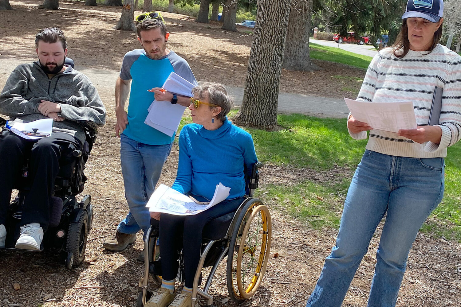 Four adults gather outdoors in a park with trees. Two people use wheelchairs, and two are standing. They are looking at papers and appear to be having a discussion or meeting.