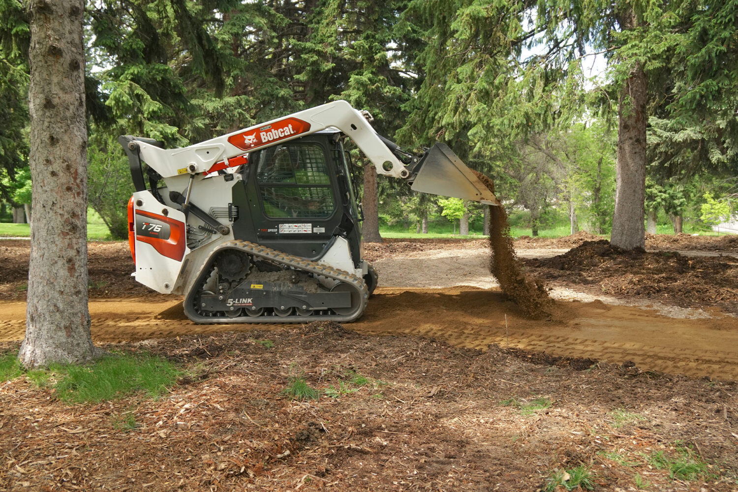 A Bobcat T76 compact track loader spreads dirt on a path in a wooded area, with tall evergreen trees surrounding the site.