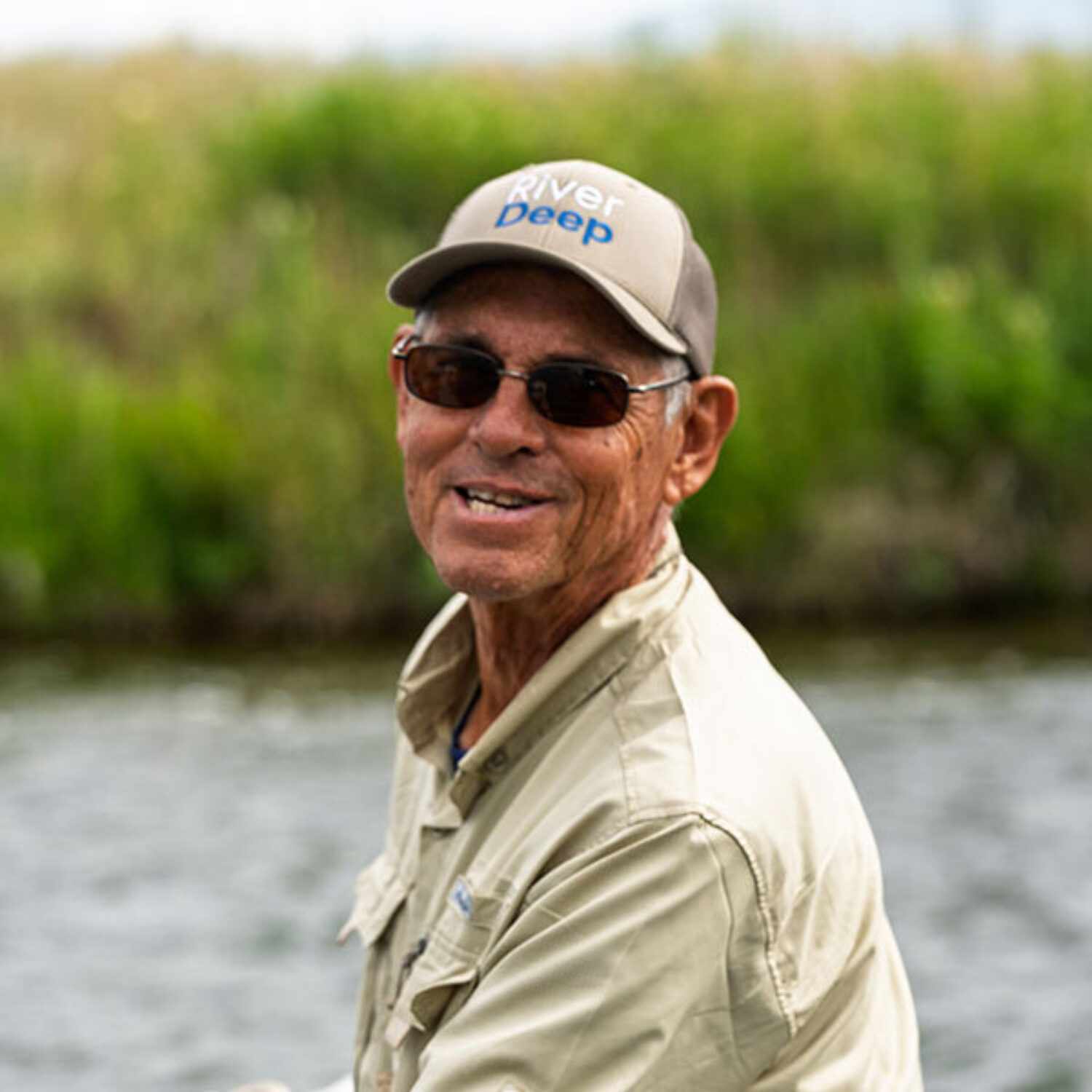 An older man wearing sunglasses and a "River Deep" cap smiles outdoors by a river, with green grass and blurred vegetation in the background.