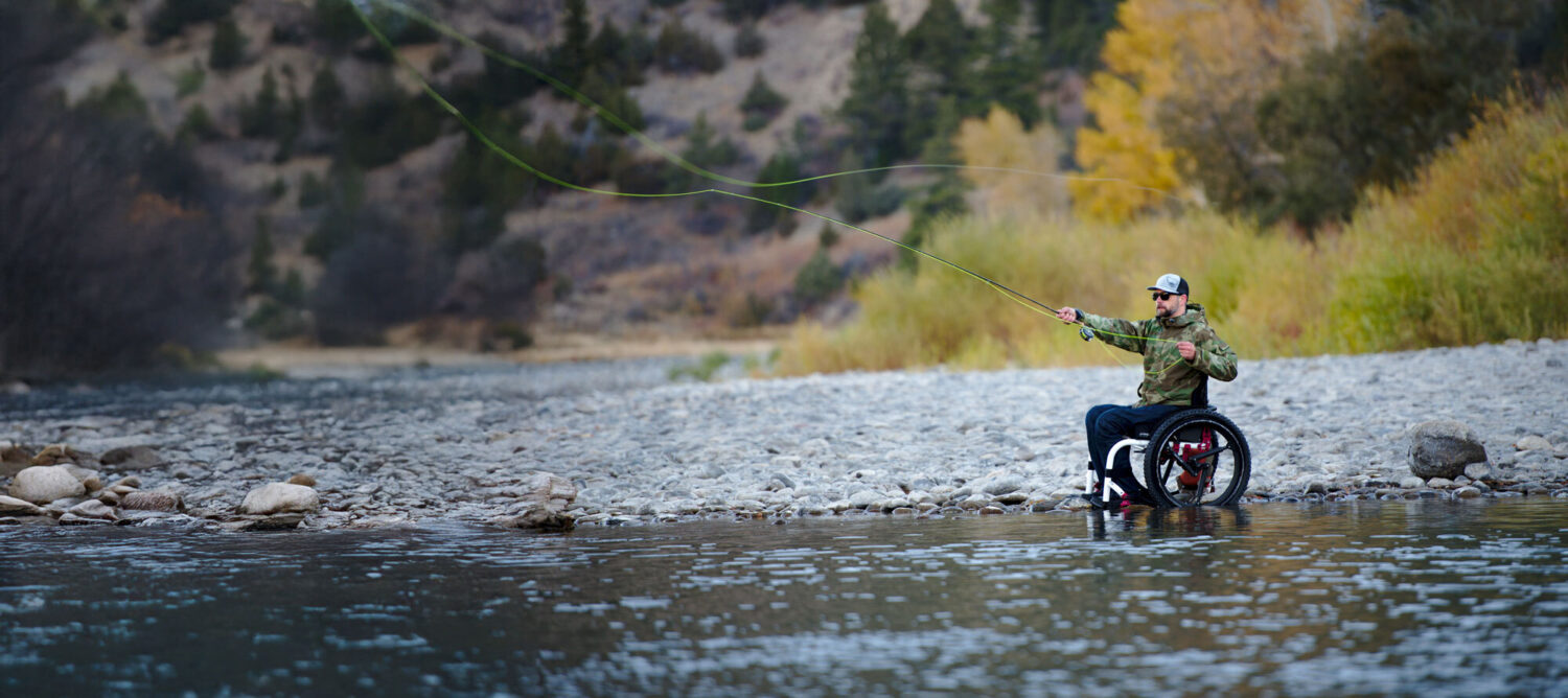  
			 A person in a wheelchair is fly fishing near the edge of a rocky riverbank, surrounded by trees and autumn foliage.
		  		 